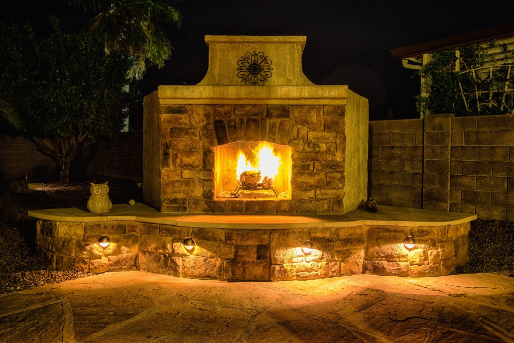Nighttime photo of DIY outdoor fireplace with stucco, stone, mantel and metal art.  Fire burning in firebox with LED lighting below hearth seating.  Flagstone surface on seating and patio in front of fireplace.