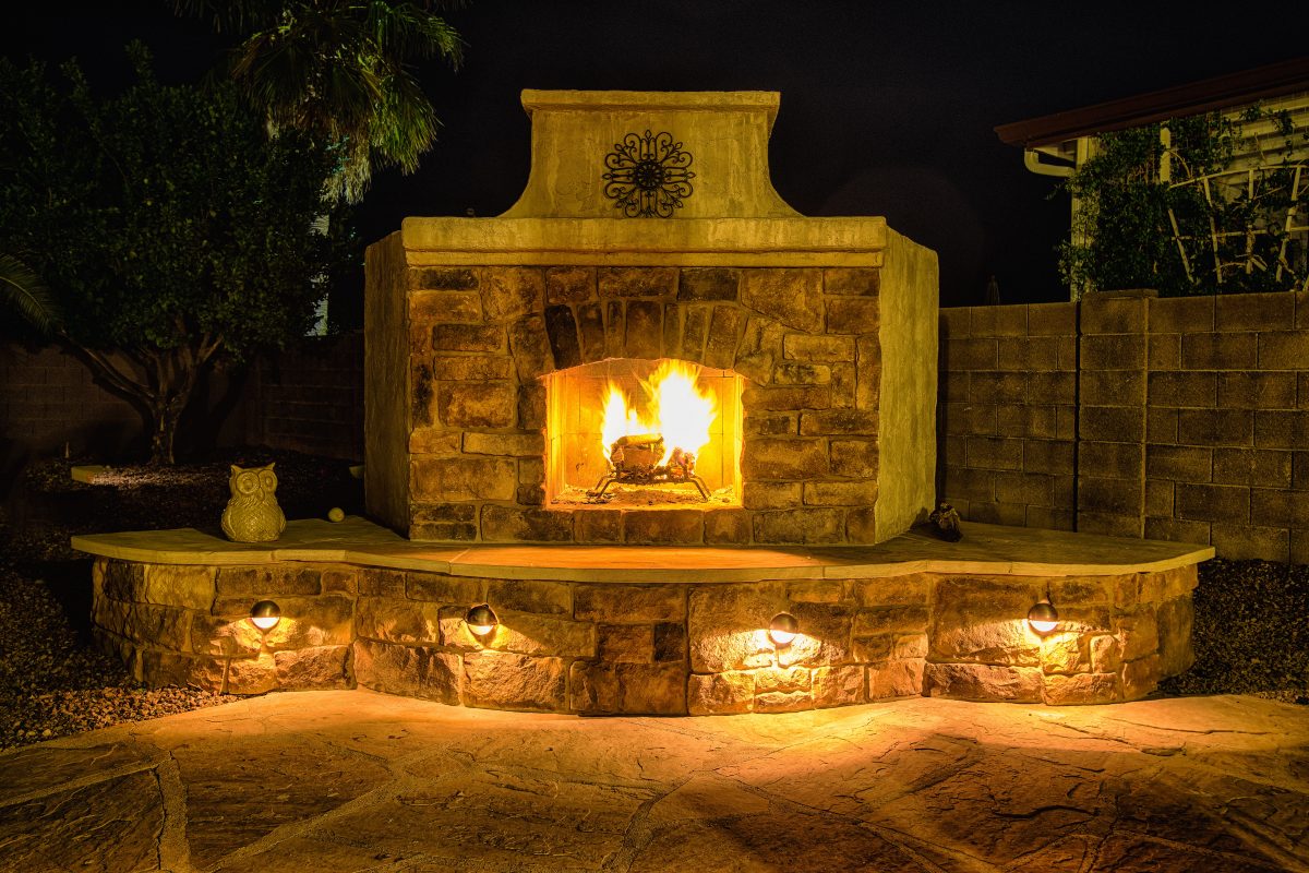 Nighttime photo of DIY outdoor fireplace with stucco, stone, mantel and metal art. Fire burning in firebox with LED lighting below hearth seating. Flagstone surface on seating and patio in front of fireplace.