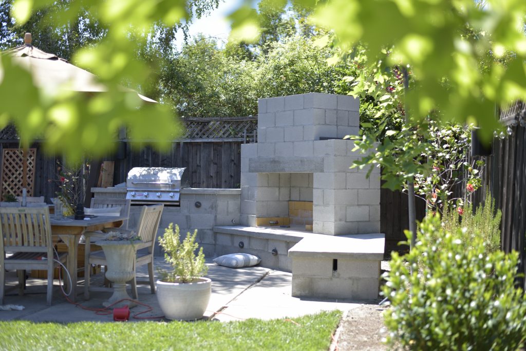 Cinder block construction outdoor fireplace and outdoor kitchen under construction in a backyard.  This fireplace is surrounded by chairs and is under trees.