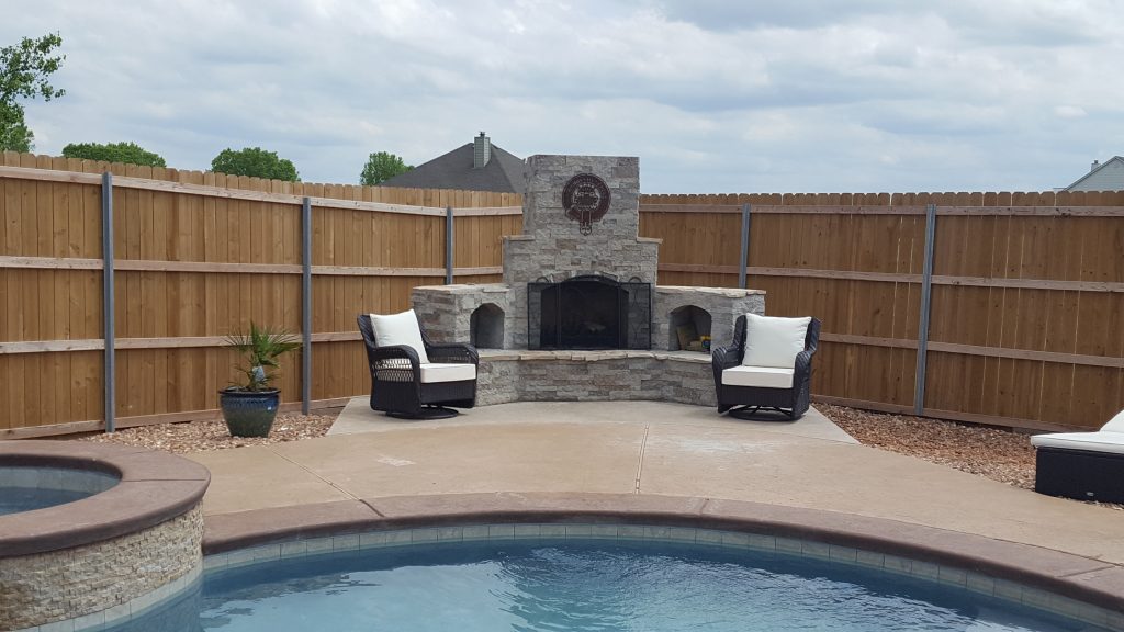 DIY outdoor fireplace built from cinderblocks and mortar covered in stone veneer. Metal art on chimney and pool and hot tub in front of fireplace. Patio furniture and wooden fence. Construction zone in backyard of Cholla design.