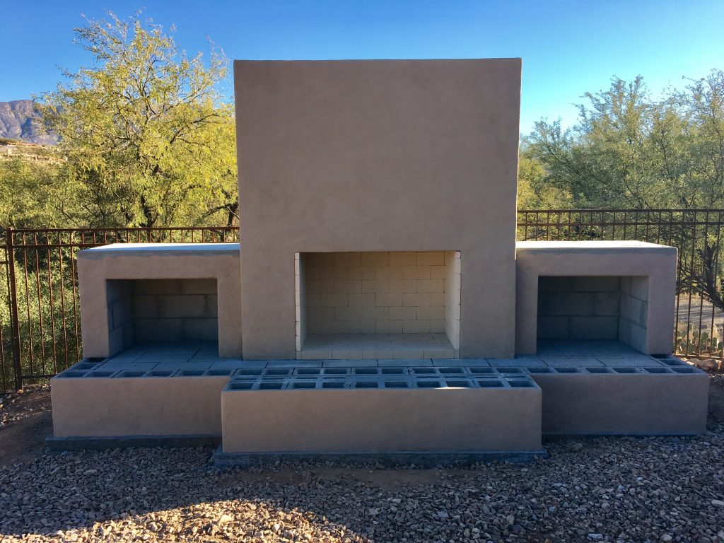 DIY outdoor fireplace built from cinderblocks and mortar covered in stucco and paint.  Construction zone in backyard of Santa Cruz design with desert foliage behind and mountains.