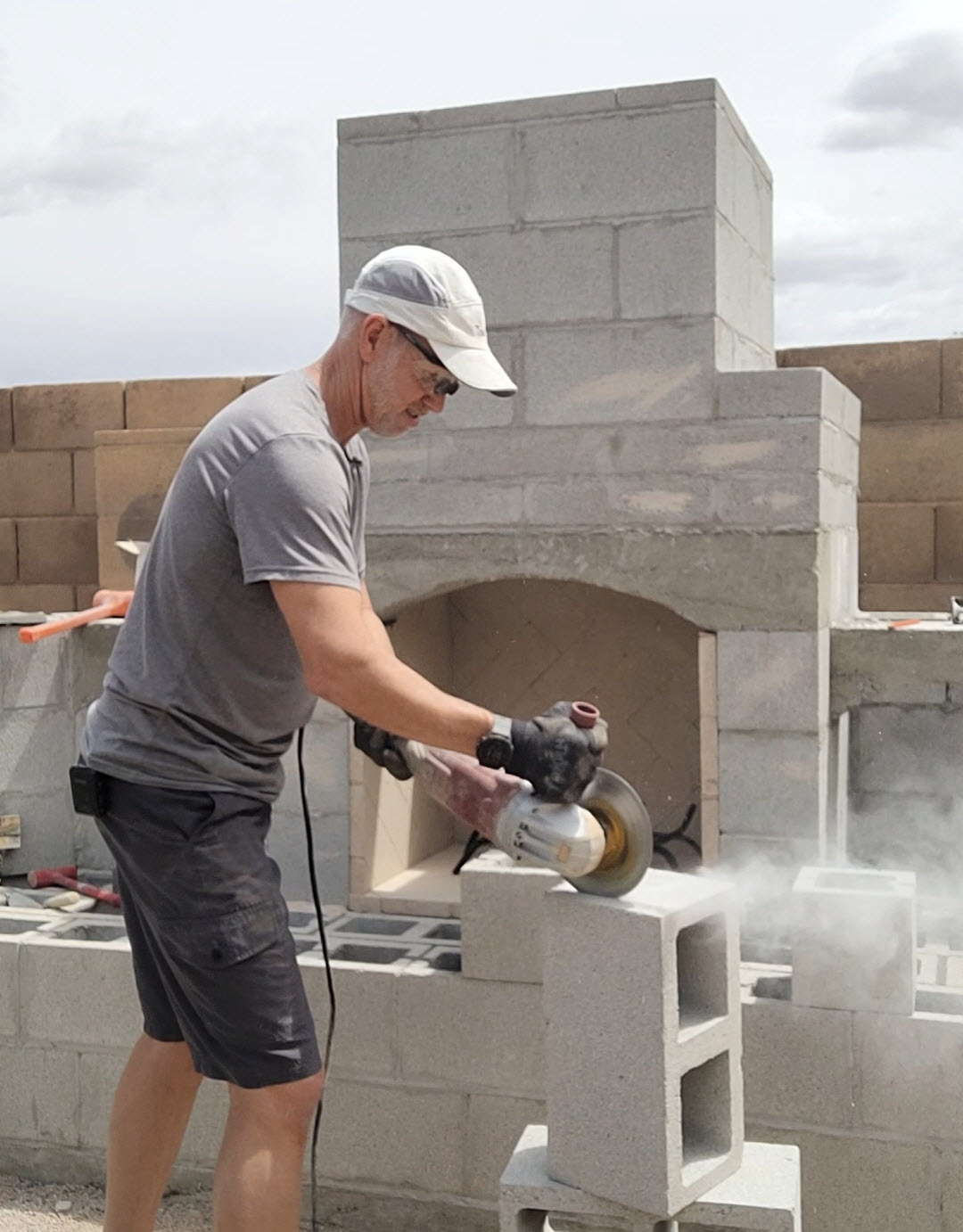 Man in white hat cutting cinderblock. Masonry DIY outdoor fireplace behind him. Diamond blade and grinder being used on construction.