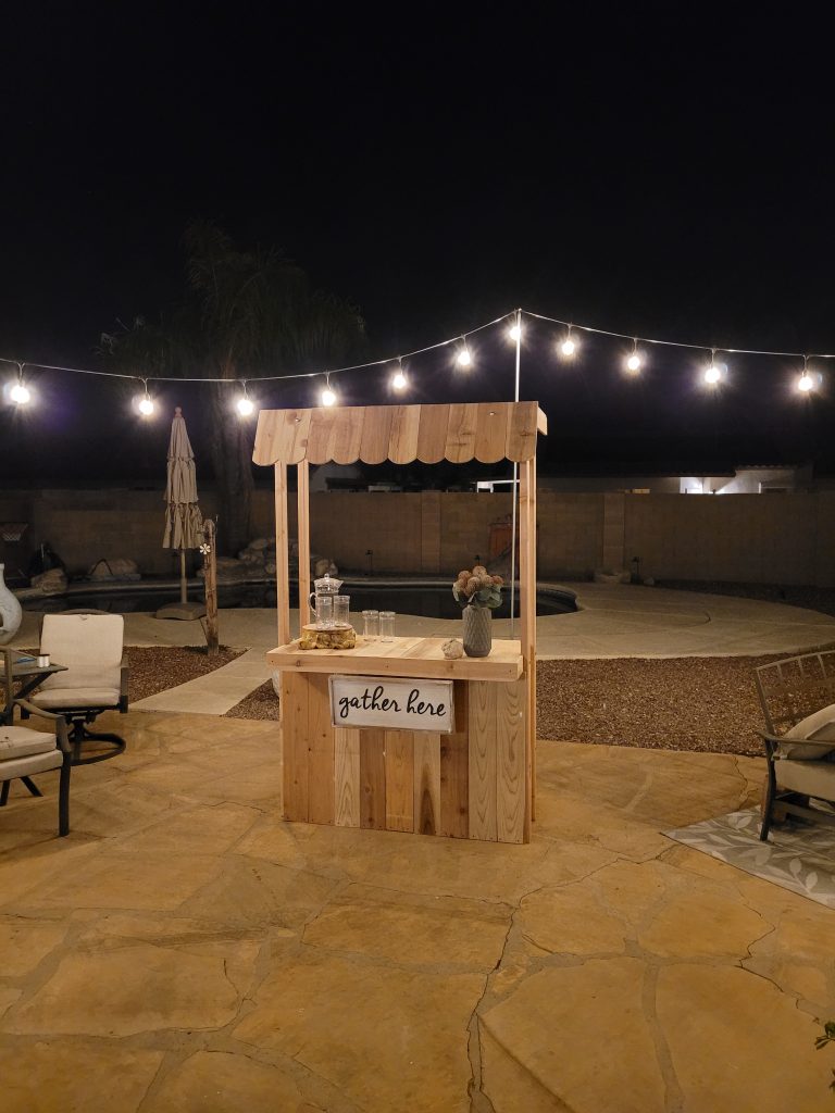 Wooden beverage bar, hot chocolate stand, lemonade stand, built in backyard.  Decorated with vase, flowers, pitcher and glasses. Swimming pool and outdoor fireplace in background.