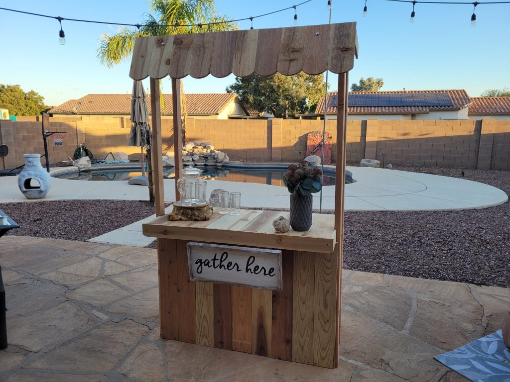 Wooden beverage bar, hot chocolate stand, lemonade stand, built in backyard.  Decorated with vase, flowers, pitcher and glasses. Swimming pool and outdoor fireplace in background.