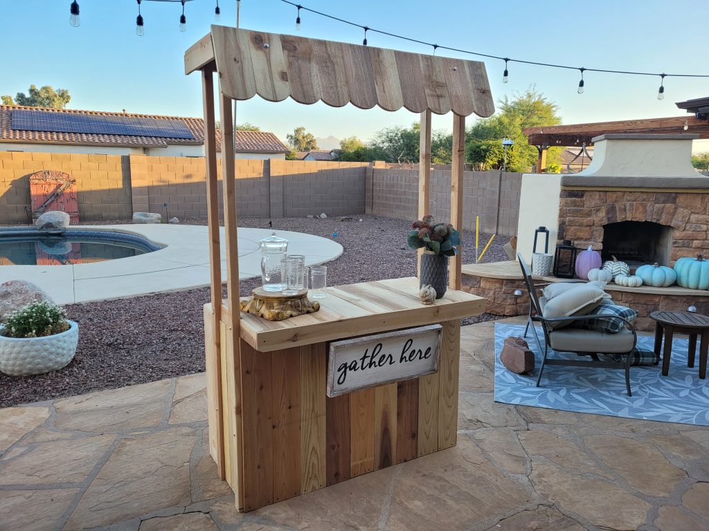 Wooden beverage bar, hot chocolate stand, lemonade stand, built in backyard. Decorated with vase, flowers, pitcher and glasses. Swimming pool and outdoor fireplace in background.