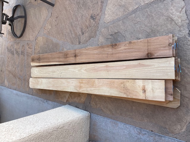 Wood laying in a stacked fashion on a flagstone patio next to a house patio post.  Outdoor furniture is next to the wooden fence slats.