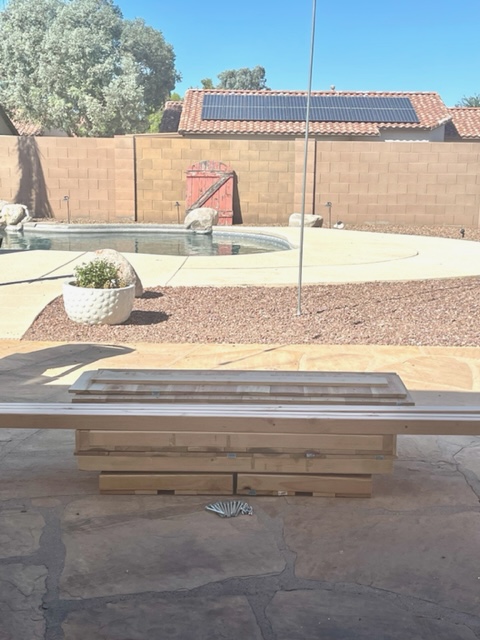 Wooden beverage bar, hot chocolate stand, lemonade stand, built in backyard.  Decorated with vase, flowers, pitcher and glasses. Swimming pool and outdoor fireplace in background.