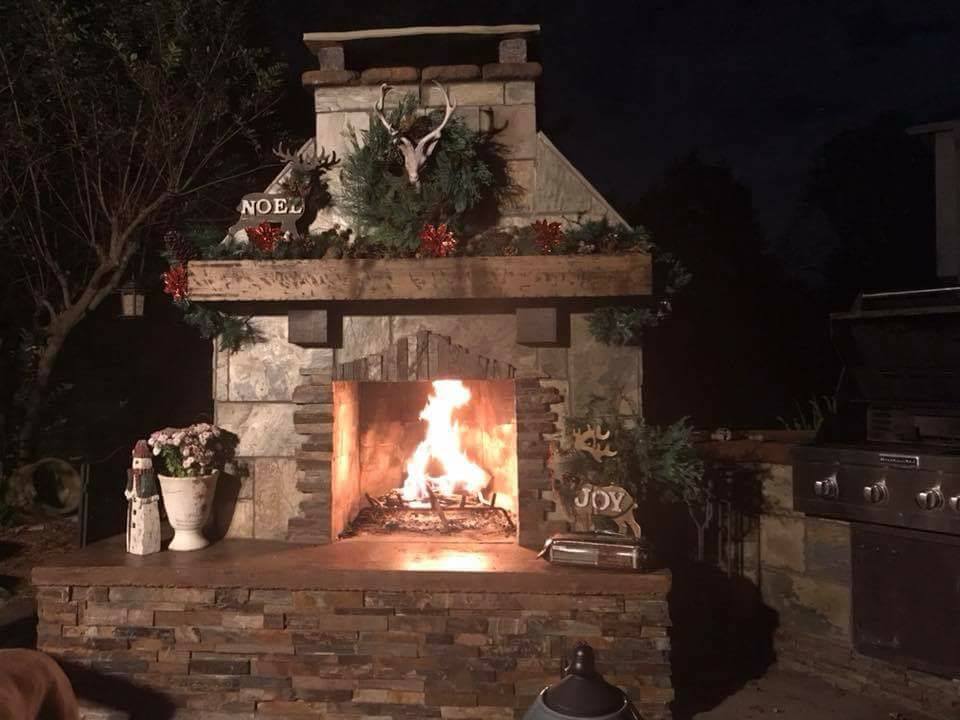 Christmas Fireplace with fire blazing. Veneer decoration with wood mantel and seating. Vases and pots with flowers and chimney cap stone.